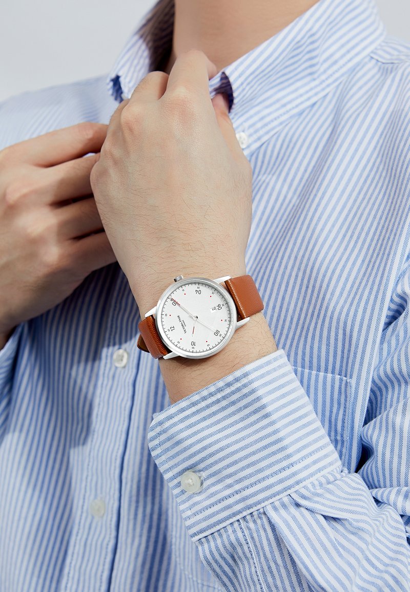 Montre à bracelet en cuir brun avec un cadran blanc, un boîtier en argent et des index noirs, portée sur une main vêtue d'une chemise rayée bleu clair.