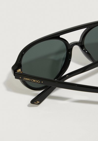 Melni saulesbrilles ar tumšiem stikliem, kas aprīkotas ar spīdīgu plastikāta rāmi, zelta akcentiem uz tempļiem un zīmola nosaukumu "JIMMY CHOO" iegravētu.