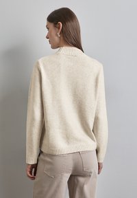 Pull en beige clair en tissu doux, avec un col et des poignets côtelés, à la coupe décontractée, porté sur un pantalon de couleur claire.