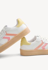 Paire de baskets blanches avec des accents jaunes pâles au talon, des bandes latérales roses, des semelles beiges texturées et des lacets blancs sur fond blanc.