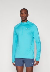 Uomo che indossa un top da corsa Nike a maniche lunghe turchese con zip a un quarto e pantaloni corti Nike blu navy, in piedi contro uno sfondo chiaro e semplice.