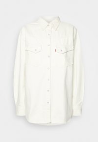 Camisa de mezclilla blanca de manga larga con botones a presión, cuello puntiagudo, bolsillos de solapa en el pecho y una pequeña etiqueta de logo roja en el bolsillo izquierdo.