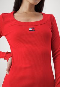 Femme portant un haut rouge côtelé ajusté à manches longues, avec un col carré et un petit logo "Tommy Jeans" au centre de la poitrine.