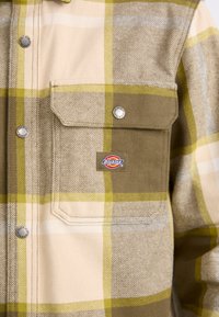 Chemise à boutons dans un motif à carreaux comprenant des teintes de beige, vert et marron. Deux poches avant et un patch logo Dickies. Matière en coton.