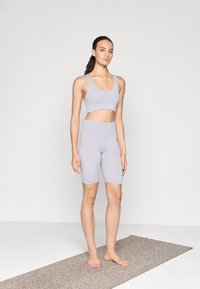 Conjunto de sujetador deportivo en gris claro y pantalones cortos de ciclismo, fabricado con un material elástico, de textura suave y diseño ajustado. De pie sobre una esterilla texturizada.