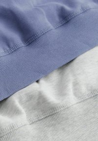 Felpa di cotone a strati in lavanda e grigio chiaro. Presenta orli a coste e texture lisce, con un aspetto leggermente stropicciato.