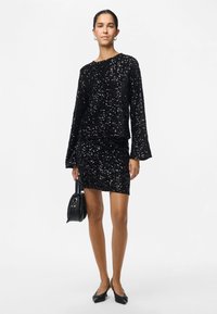 Moteris vilki juodą ilgomis rankovėmis blizgučių viršutinę dalį ir derančią blizgučių mini sijoną, laiko juodą rankinę ir avima juodus aukštakulnius batus.