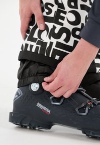 Zwarte skiboot met een gestructureerd ontwerp en verstelbare hardware. Een hand past de omslag aan van zwarte en witte gestructureerde skibroeken.