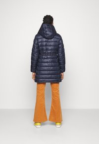 Marinblå pufferjacka med huva, med horisontell sömnad, avsmalnande midja och en slät, glänsande yta. Kombinerad med orange kordflåsade byxor med utsvängda ben.