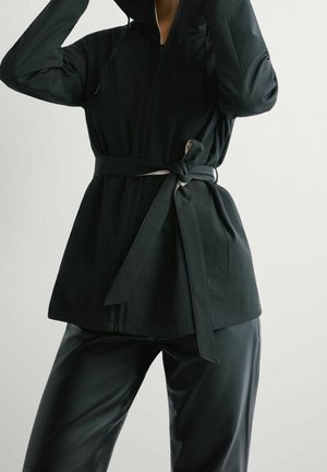 Personne portant une veste noire zippée avec une ceinture nouée à la taille et un pantalon en cuir noir, ajustant la capuche de la veste.