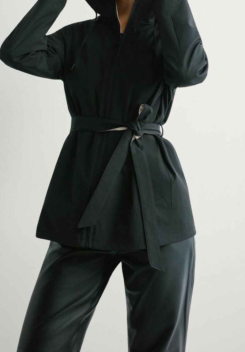 Personne portant une veste noire zippée avec une ceinture nouée à la taille et un pantalon en cuir noir, ajustant la capuche de la veste.