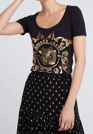 Femme portant un t-shirt noir à manches courtes avec le logo doré Versace Jeans et une jupe noire à pois dorés sur un fond uni.