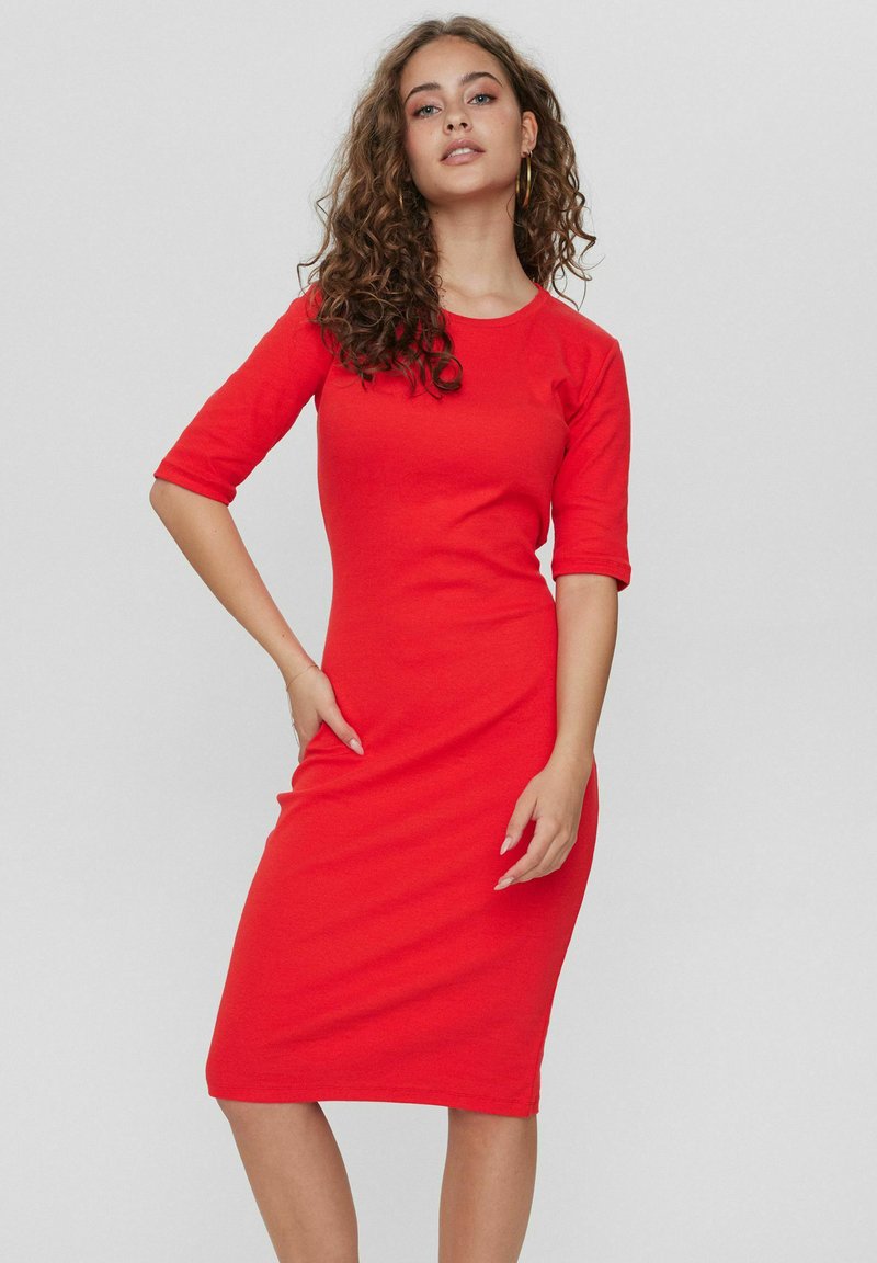 Abito fitted rosso realizzato in tessuto morbido, con collo rotondo e maniche a tre quarti, che termina appena sotto il ginocchio con una texture liscia.