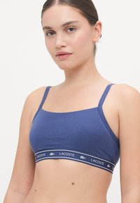 Blå bomull bralette med tynne stropper og en bred merkebånd med Lacoste-logo i hvitt. Glatt tekstur, tettsittende design.