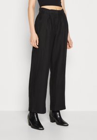 Široké černé kalhoty z texturované látky s záhyby vpředu, spárované s černými kotníkovými botami s krokodýlím vzorem.