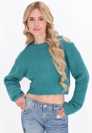 Gekürzter gestrickter Pullover in Teal mit geripptem Muster, mit verlängerten Ärmeln und einem Schulterausschnitt. Kombiniert mit hellblauen Jeans.