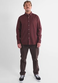 Chemise à manches longues bordeaux à boutons avec col, associée à un pantalon marron et des baskets noires avec des accents blancs.