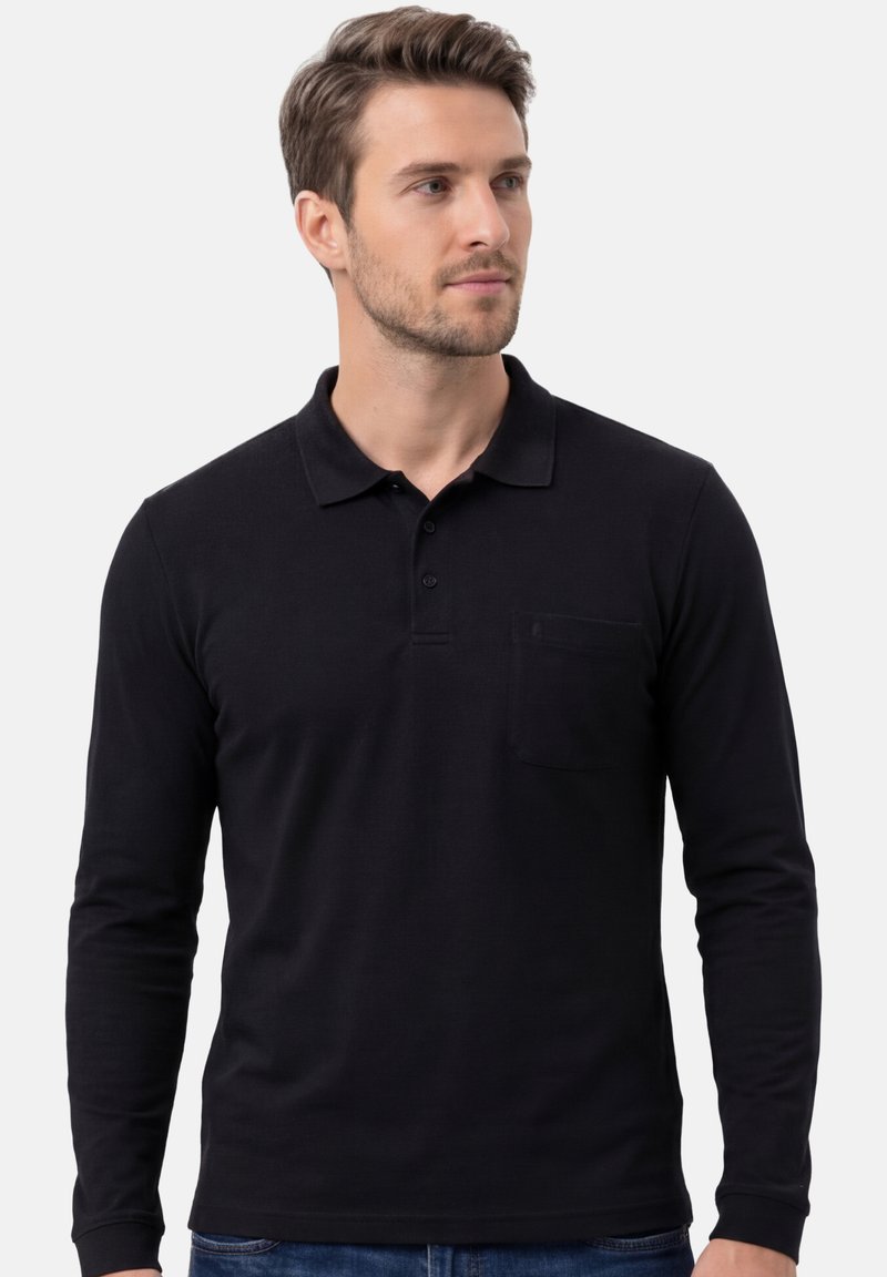 Homme portant un polo noir à manches longues avec poche, debout devant un fond clair uni, regardant sur le côté.