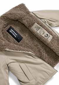 Giacca isolante beige con fodera in pelliccia sintetica marrone, dotata di chiusura a zip, tasche laterali e etichette per la cura del tessuto. Impermeabile e antivento.
