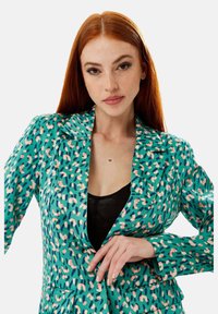 Green blazer with a pattern of pink and navy shapes. Features a notched lapel and two front pockets, showcasing a smooth texture.