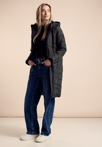 Gevoerde zwarte lange jas met capuchon, gedragen over een zwart topje en blauwe wijde spijkerbroek, gecombineerd met witte sneakers. Glad textuur en gestructureerd ontwerp.