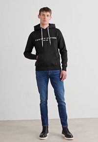 Juodas džemperis su balta "Tommy Hilfiger" logotipu ir virvutėmis, derinamas su mėlynomis džinsinėmis kelnėmis ir juodais aukštakulniais sportbačiais. Balti pado akcentai.