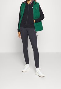 Gilet imbottito verde sopra una felpa nera con zip, abbinato a leggings neri e sneakers bianche. Presenta loghi e accenti bicolore.