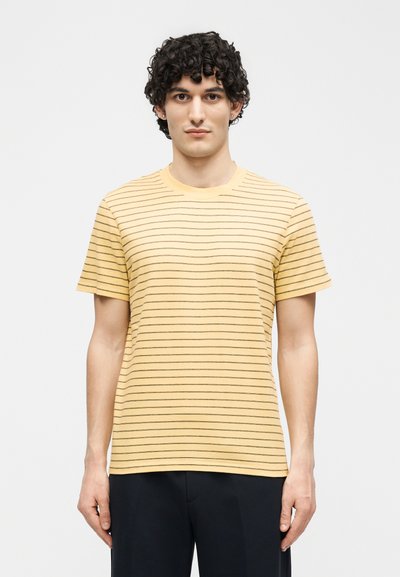 Νέος άνδρας με σγουρά σκούρα μαλλιά που φοράει κίτρινο κοντομάνικο μπλουζάκι με λεπτές μαύρες οριζόντιες ρίγες και σκούρο παντελόνι, όρθιος μπροστά σε λευκό φόντο.