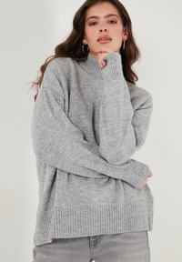 Vaaleanharmaa neulepaita, jossa on korkea kaulus, ribatut hihansuut ja helma. Yliompelumuotoinen malli, jossa on roikkuvat hartiat ja tekstuuri.
