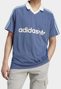 Blå atletisk tröja med vita vertikala ränder, en vit krage och "adidas"-logotyp i vitt. Tillverkad av ett lätt material.