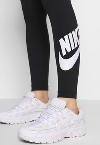 Zwarte leggings met groot wit "NIKE" logo aan de zijkant, gecombineerd met witte sportschoenen met geperforeerde details en een contrasterende zool.