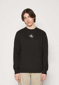 Black crewneck sweatshirt made from cotton, featuring a white Calvin Klein logo at the chest. Simple design with ribbed cuffs and hem.