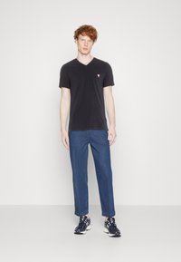 Sort V-hals t-shirt med et lille rødt trekantet logo, parret med blå denimbukser og sorte sneakers med hvide detaljer.
