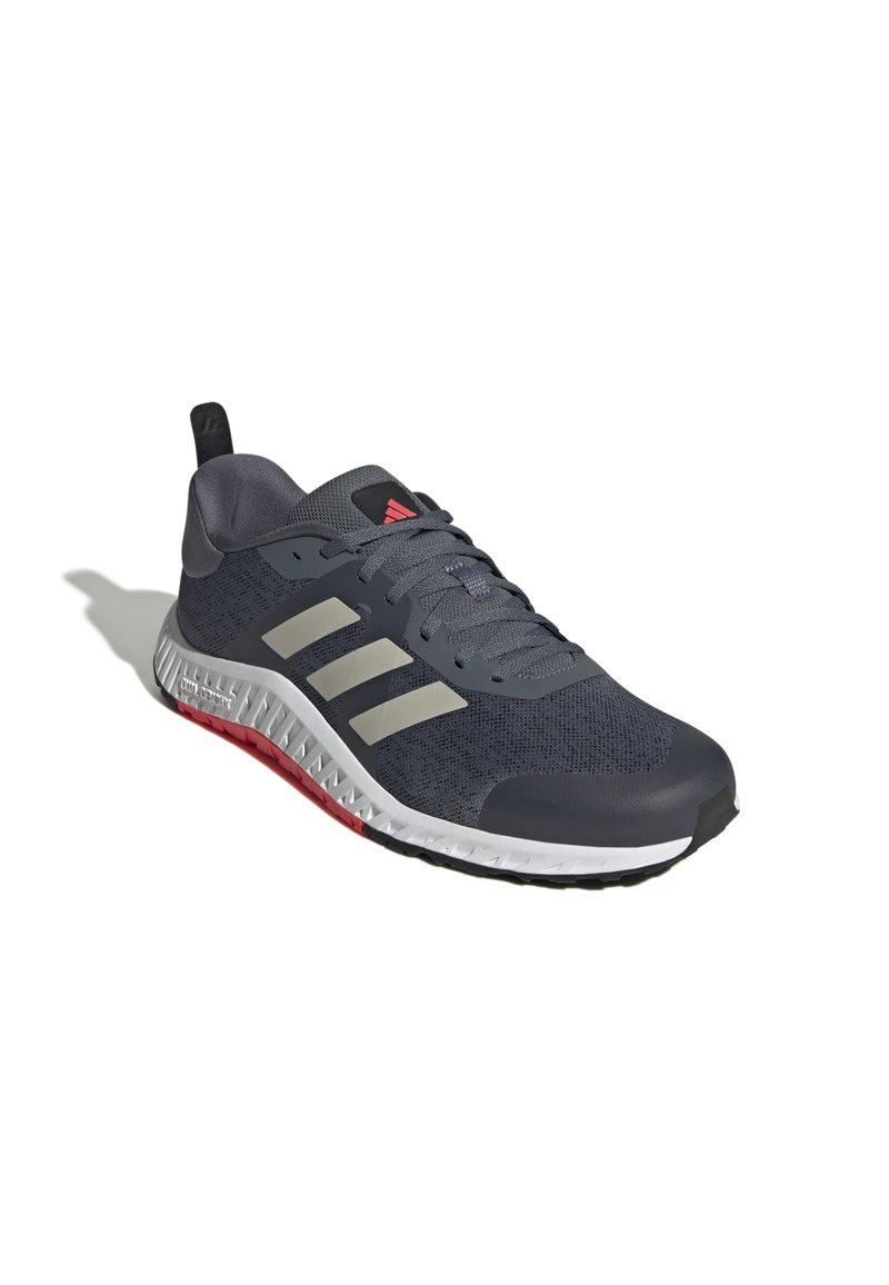 Zapato deportivo para hombres en malla gris oscuro con tres rayas gris claro. Presenta acentos rojos en la suela y una entresuela blanca texturizada.