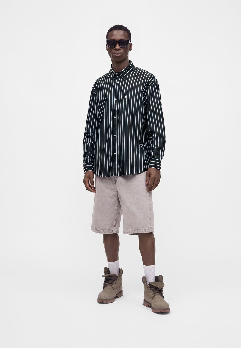 Mann trägt ein schwarz-weiß vertikal gestreiftes Langarmhemd, hellgraue Shorts, beige Stiefel, weiße Socken und schwarze Sonnenbrille, steht vor weißem Hintergrund.