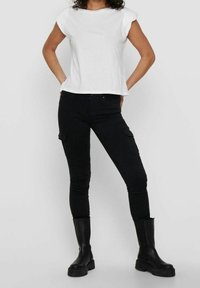 Top corto de manga corta blanca, combinado con pantalones cargo negros ajustados que cuentan con bolsillos laterales. La modelo lleva botas negras hasta la rodilla. Fondo liso.