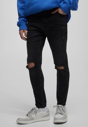 Personne portant un jean slim noir déchiré, des baskets blanches et un sweat-shirt bleu avec une main dans la poche sur fond uni.
