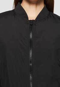 Veste bomber noire avec col montant, fermeture éclair à l'avant et tissu lisse et texturé. Comprend des poignets et une ceinture élastiqués.