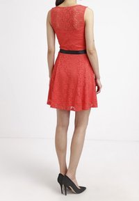 Robe en dentelle rouge avec un corsage ajusté et une jupe évasée, dotée d'une ceinture noire à la taille et d'une finition texturée. Design à dos ouvert. Talons noirs.