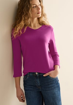 Fuchsia Langarmoberteil aus einem weichen Stoff, mit rundem Halsausschnitt und lockerem Schnitt, kombiniert mit dunkelblauen Jeans.