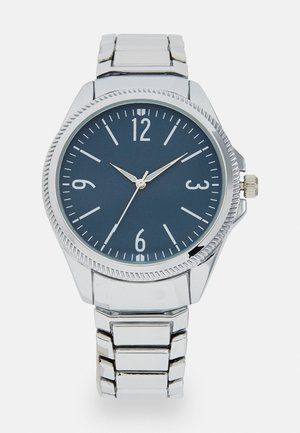Sølv rustfritt stålklokke med en mørkeblå rund urskive. Har hvite timemarkører og visere, teksturert bezel og justerbar lenke.