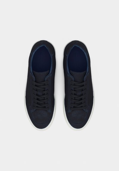 Ζευγάρι μαύρα αθλητικά παπούτσια χαμηλού ύψους με λευκές σόλες και μαύρα κορδόνια, απαθανατισμένα από πάνω σε απλό λευκό φόντο.