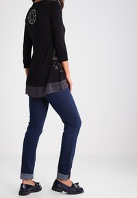 Svart långärmad topp med dekorativa mönster på baksidan och en texturerad grå nederkant, ihop med mörka jeans och svarta skor.