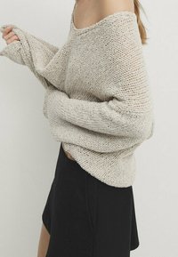 Beige, off-the-shoulder knitted sweater with loose, wide sleeves, paired with a black mini skirt, showcasing a relaxed silhouette and texture.