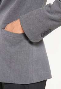 Šedý blazer s texturovanou tkaninou, který má jedno přední kapsu a tři tmavé knoflíky na manžetě. Přiléhavý střih a rovný lem.