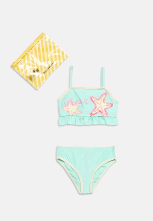 Costume da bagno per ragazza a due pezzi di colore azzurro chiaro con disegni di stelle marine rosa e rifinitura con balze, accanto a una borsa di confezione gialla a righe.