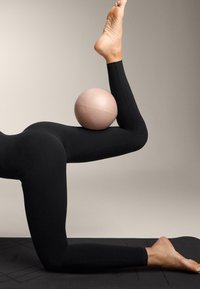 Fekete nadrágot viselő személy egy rózsaszín tornalabdát tart egy hajlított vádli és comb között, miközben térdel egy fekete szőnyegen.