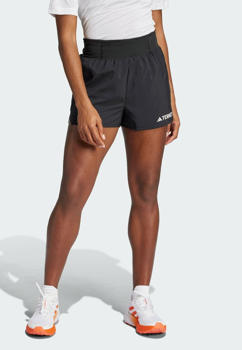 Shorts atléticos negros con una cintura ancha, que presentan el texto "TERREX" y un logo de Adidas. Combinados con zapatos blancos con acentos en naranja.