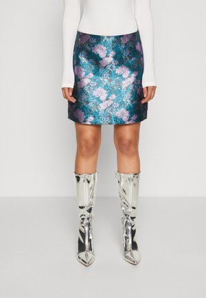 Kvinde iført en metallic blå nederdel med lavendel blomsterprint, hvid bluse med lange ærmer og skinnende sølv knæstøvler.