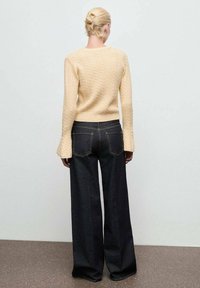 Una donna con capelli biondi raccolti in uno chignon indossa un maglione beige a trecce e jeans scuri a vita alta e gamba larga, mentre si trova su un pavimento marrone.